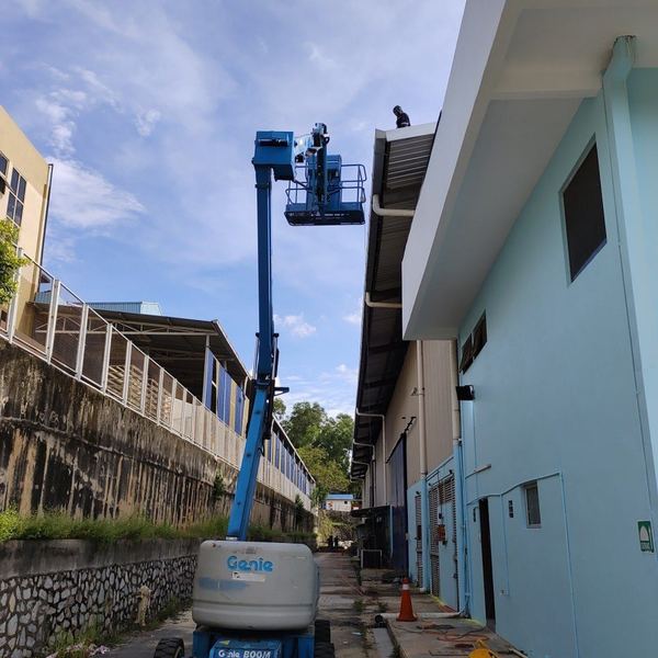 Genie boom lift raised against a warehouse wall to reach the roofline, with a worker on the roof above
