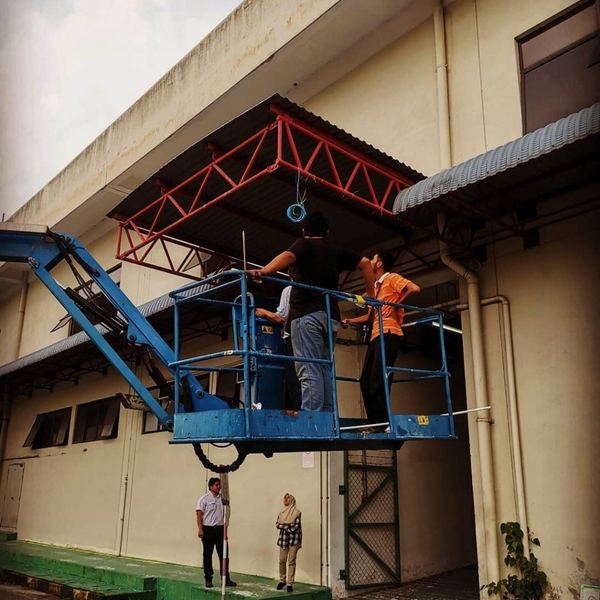 Two technicians on a raised boom lift platform running cable beneath a red steel roof truss