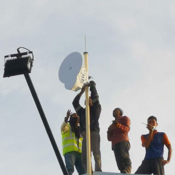 Two technicians in hi-vis vests mounting and aligning a wireless dish on a factory rooftop mast