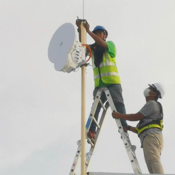 Technician on a stepladder fixing a wireless dish to a rooftop pole while a colleague steadies the ladder